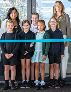 Teachers and pupils are standing behind a blue ribbon with a child holding scissors ready to cut the ribbon 