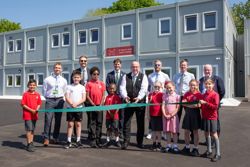 Pupils, councillors and teacher are standing in front of the school building.  A Long piece of ribbon is held both ends by pupils while the councillor cuts it 