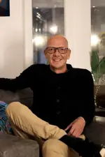 Man wearing glasses dressed in black top and khaki trousers is sat on the sofa, smiling