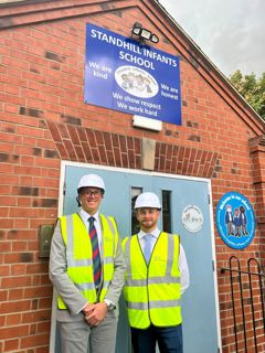 Cllrs Walker Gurley and Green are wearing hi-vis and hard hats standing outside the school entrance and sign.