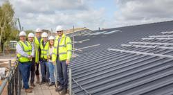 Councillor and partners are standing on a walkway by the side of the new building's roof. Everyone is wearing hard hats and hi-vis jackets