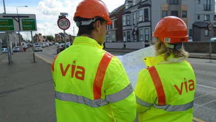 Two people wearing hi viz jackets with the Via logo on looking at drawings next to a road
